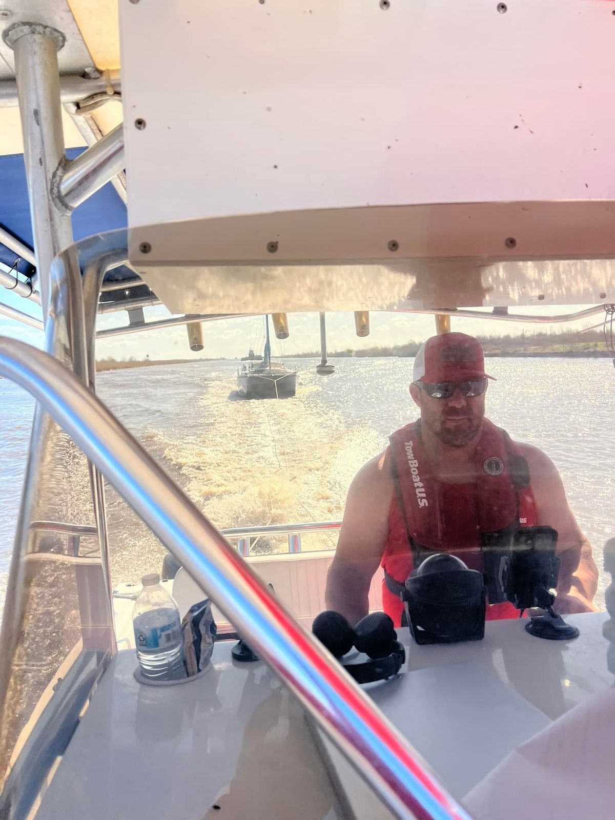 A TowBoatUS operator in a red cap and life jacket sits aboard a towing vessel on Sabine Lake, with service equipment on the deck and calm water behind.