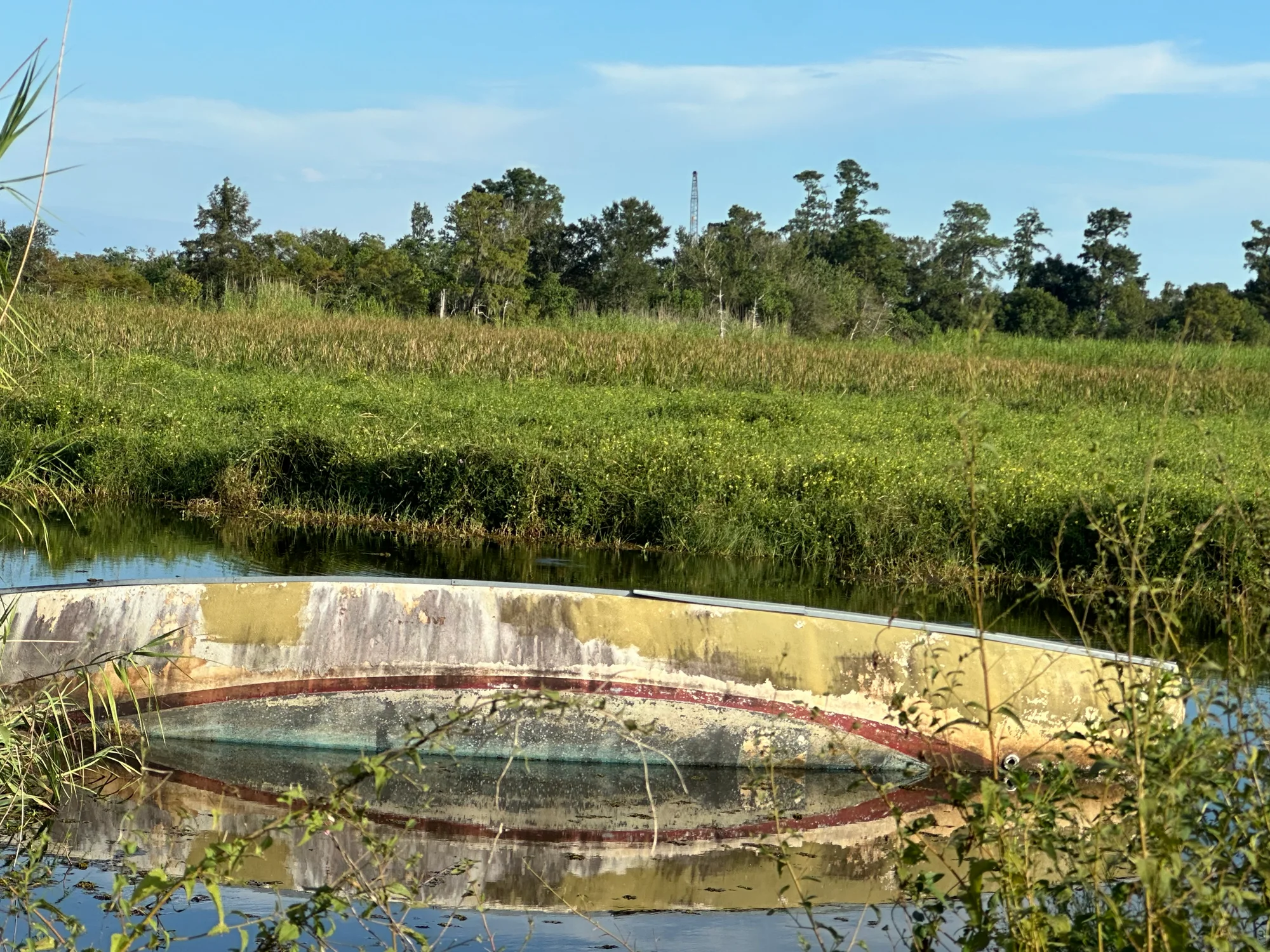 A capsized boat resting hull-up in a marsh channel near Sabine Lake, ringed by reeds and mirrored in still water.
