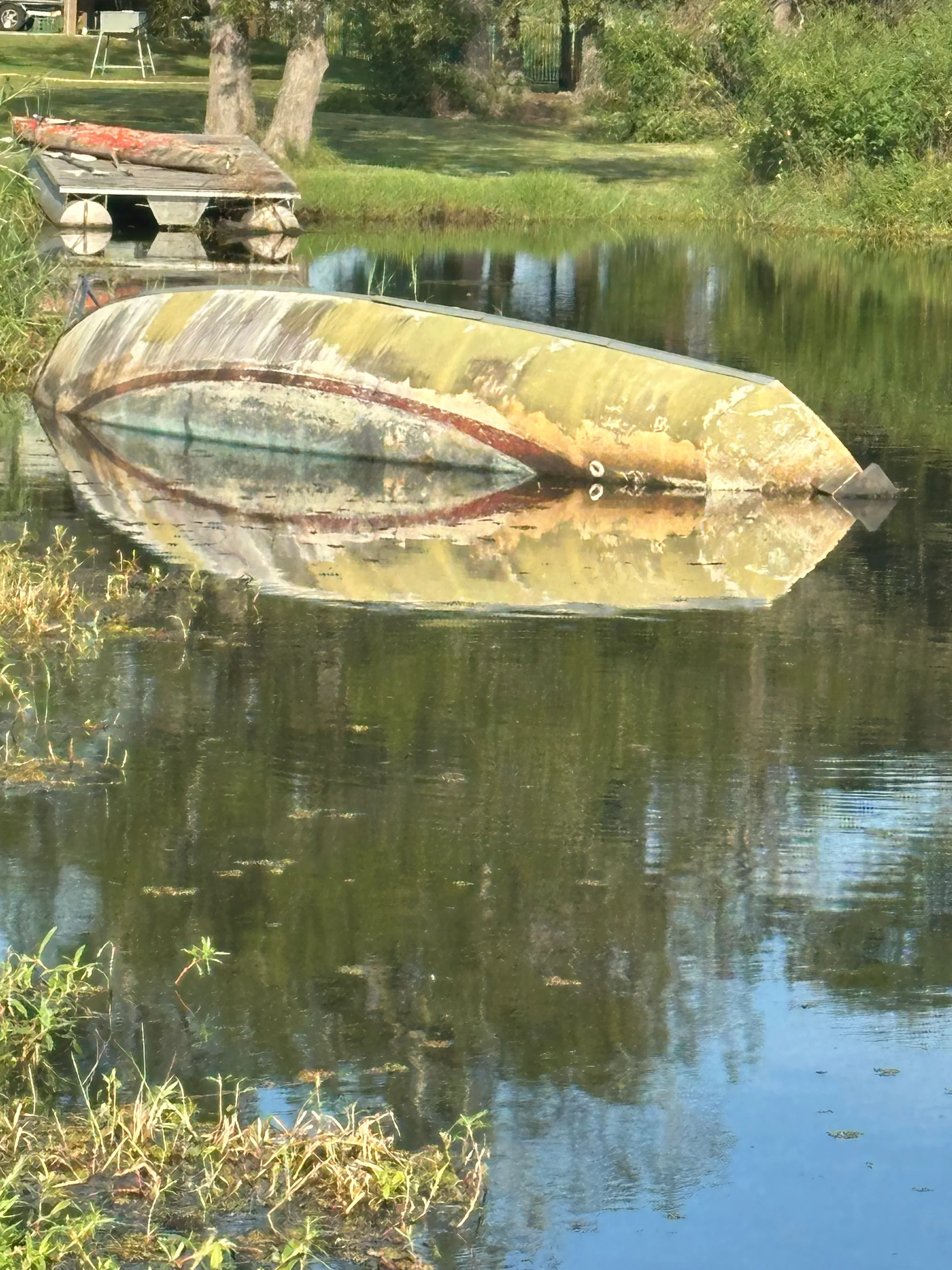 A closer view of the same capsized boat near a small dock, its hull reflected in the calm water.