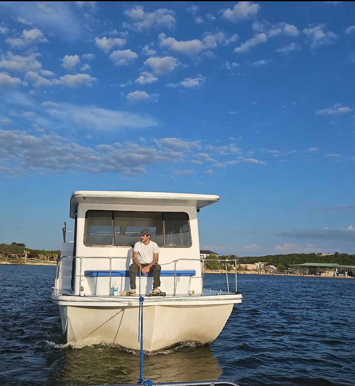 A houseboat underway on the water with a man seated on the stern deck under a clear blue sky.